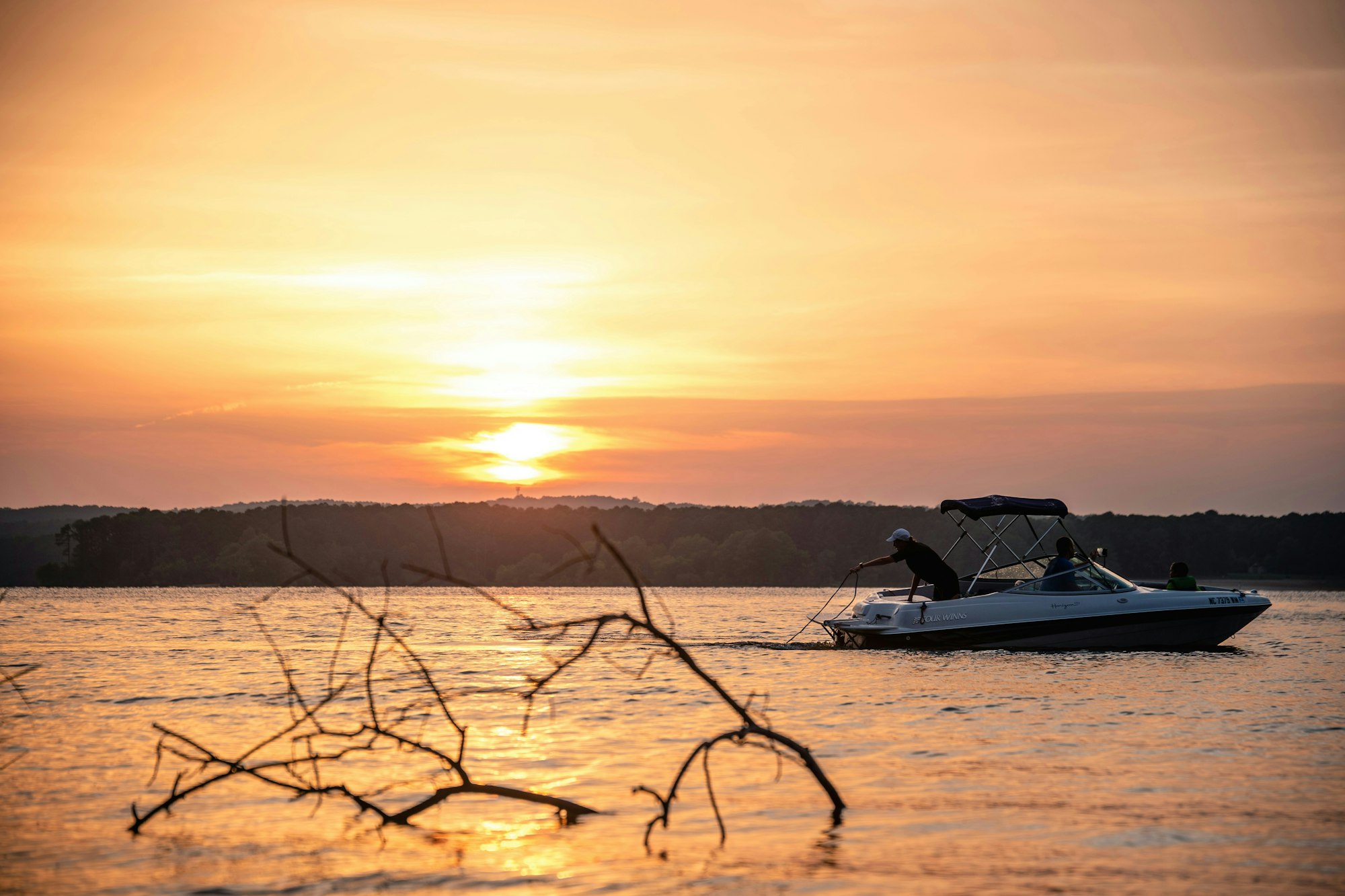 A boat with people on a lake at sunset, with a branch in the foreground and trees on the horizon. Photo by Jacob Diehl on Unsplash