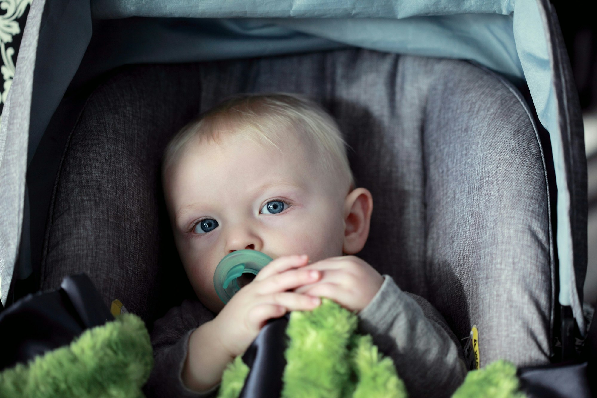 A baby with a pacifier sits in a stroller, holding a green blanket.