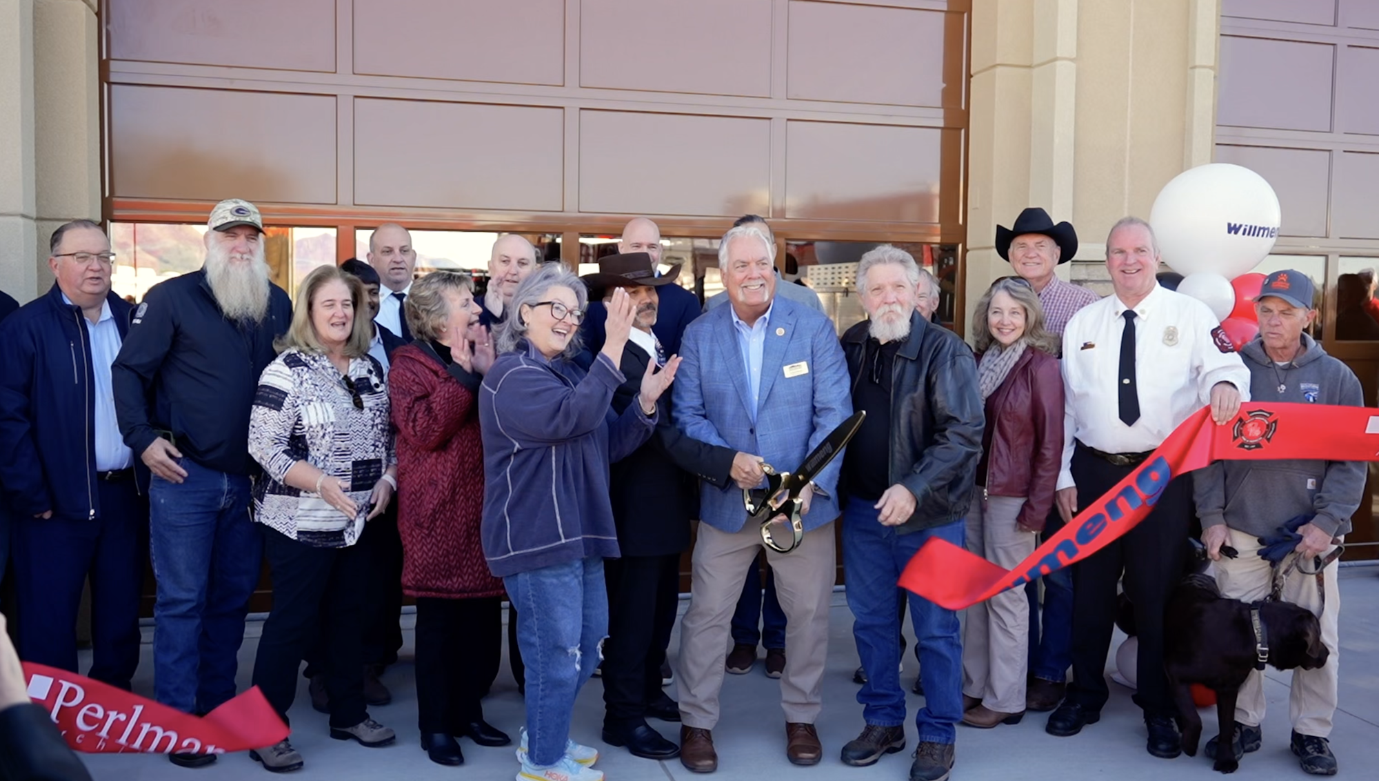 A group of people gather to celebrate a ribbon-cutting event, with one man holding scissors. Balloons and a red ribbon are visible.