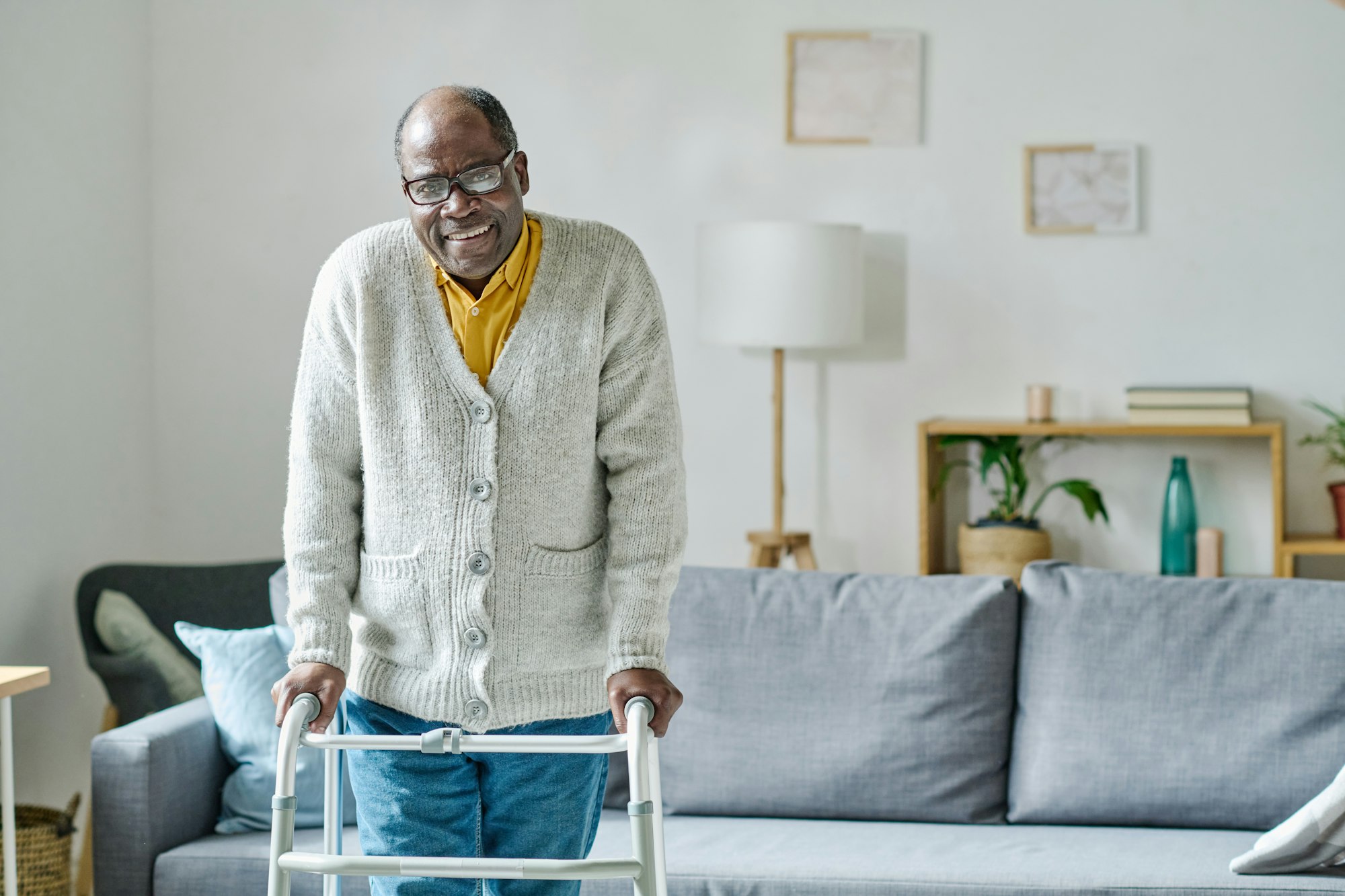 Elderly person with glasses using a walker, standing in a cozy living room with a sofa, lamp, and shelf.
