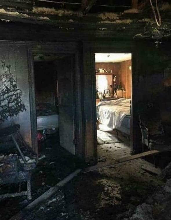 A fire-damaged room with a burnt ceiling, charred walls, debris, and an intact bedroom visible through a doorway.