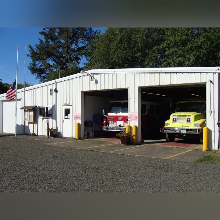 A small fire station with two fire trucks parked inside the open garage bays and an American flag outside.