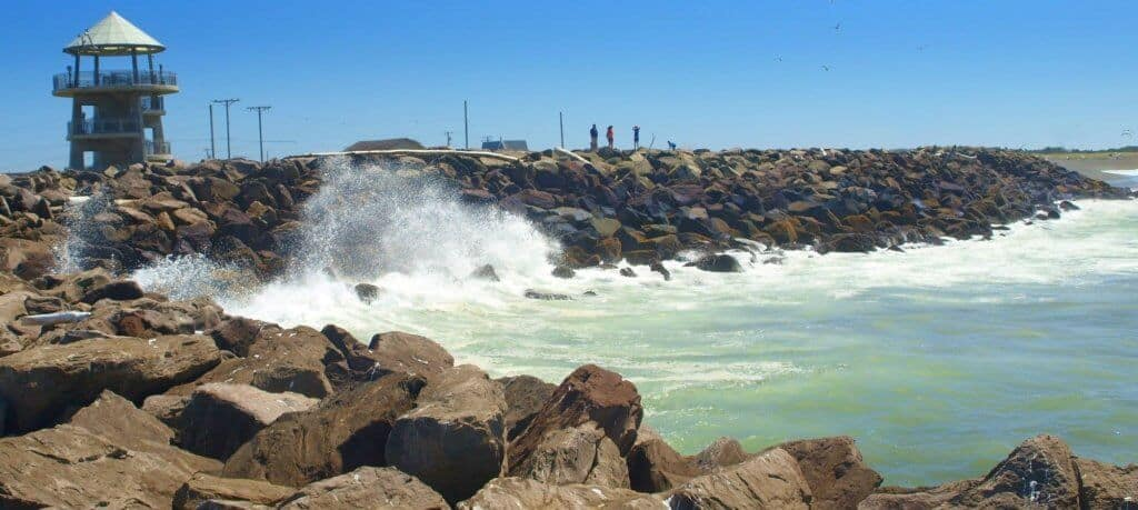 A rocky breakwater with crashing waves, a viewing tower, and people standing on the rocks under a clear blue sky.