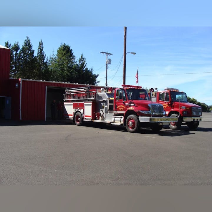 Two red fire trucks parked outside a fire station with an American flag in the background.