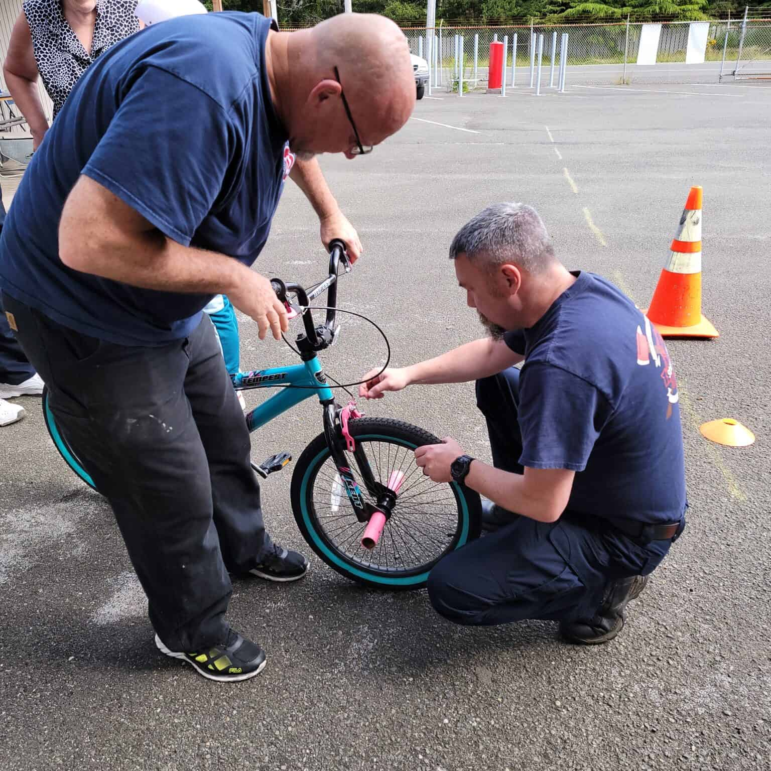 Two men are inspecting or fixing a bicycle's front wheel on an asphalt surface, with traffic cones nearby.
