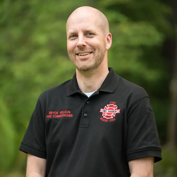 A smiling man in a black polo shirt labeled "Fire Commissioner" stands outdoors with greenery in the background.