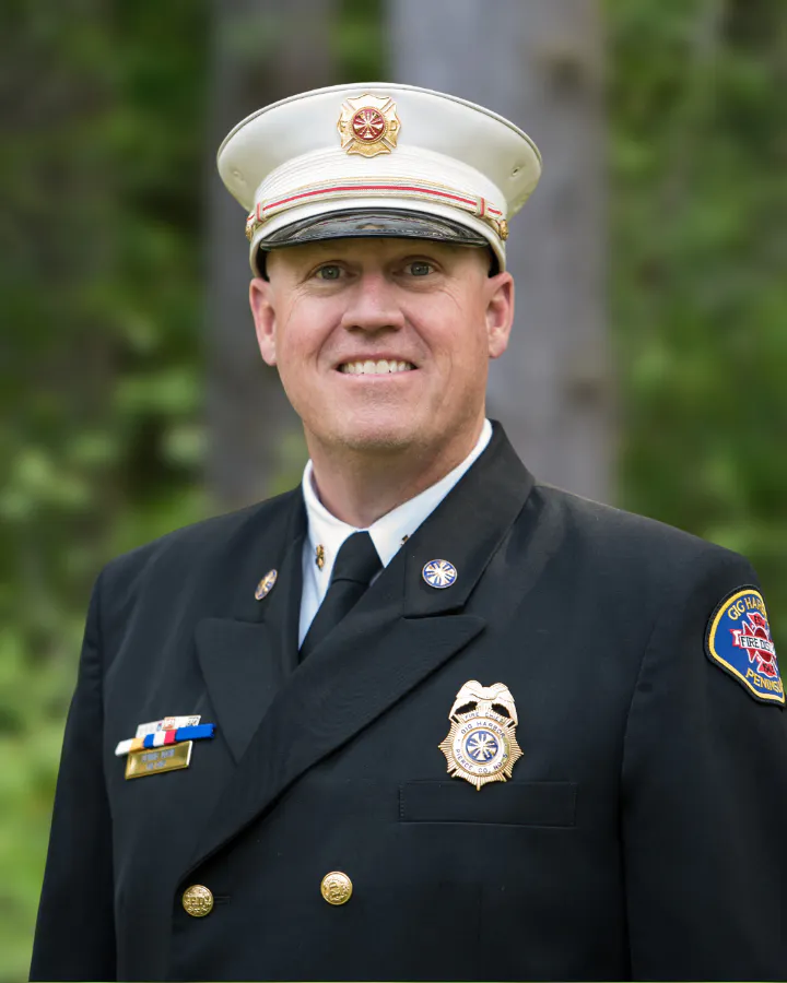 A person in a formal fire department uniform, smiling, with a distinctive hat and badges, against a green background.