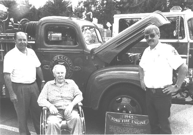Three men, one in a wheelchair, stand by a 1945 Ford fire engine, showcasing a piece of fire-fighting history.