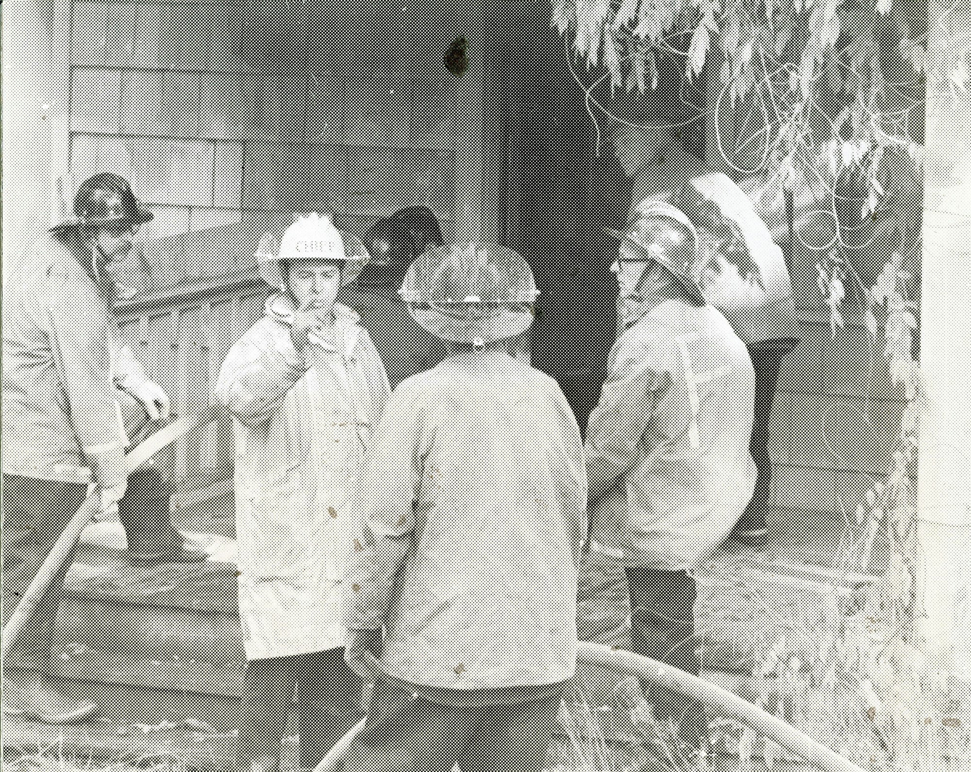 A group of firefighters in gear, discussing and preparing near a building, likely responding to an incident.