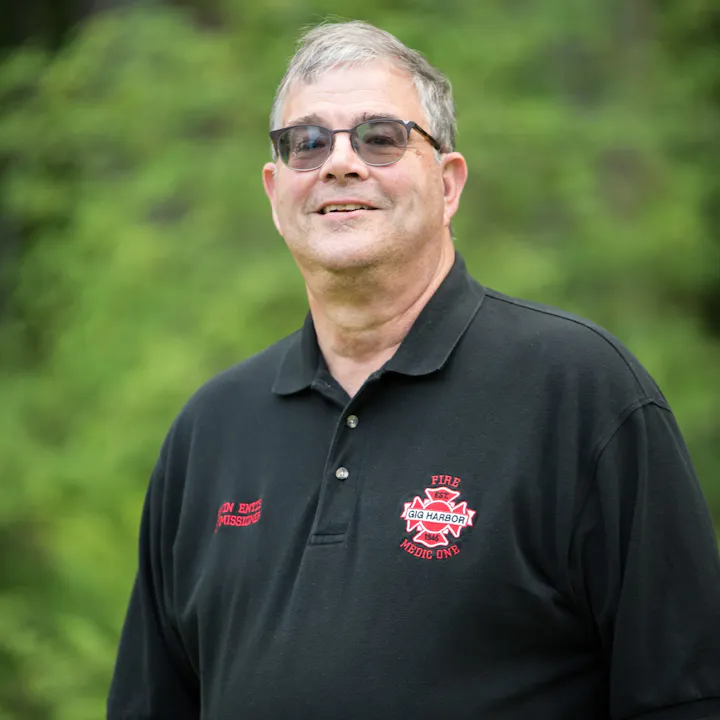 A smiling man in glasses wearing a black polo shirt with a fire and medical emblem, standing outdoors against a green background.