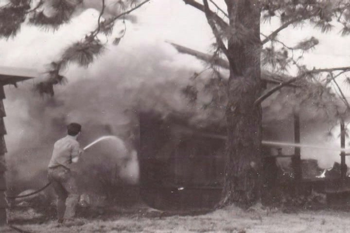 A firefighter battles a smoky blaze near a wooden structure, surrounded by trees, in this historical black-and-white photo.