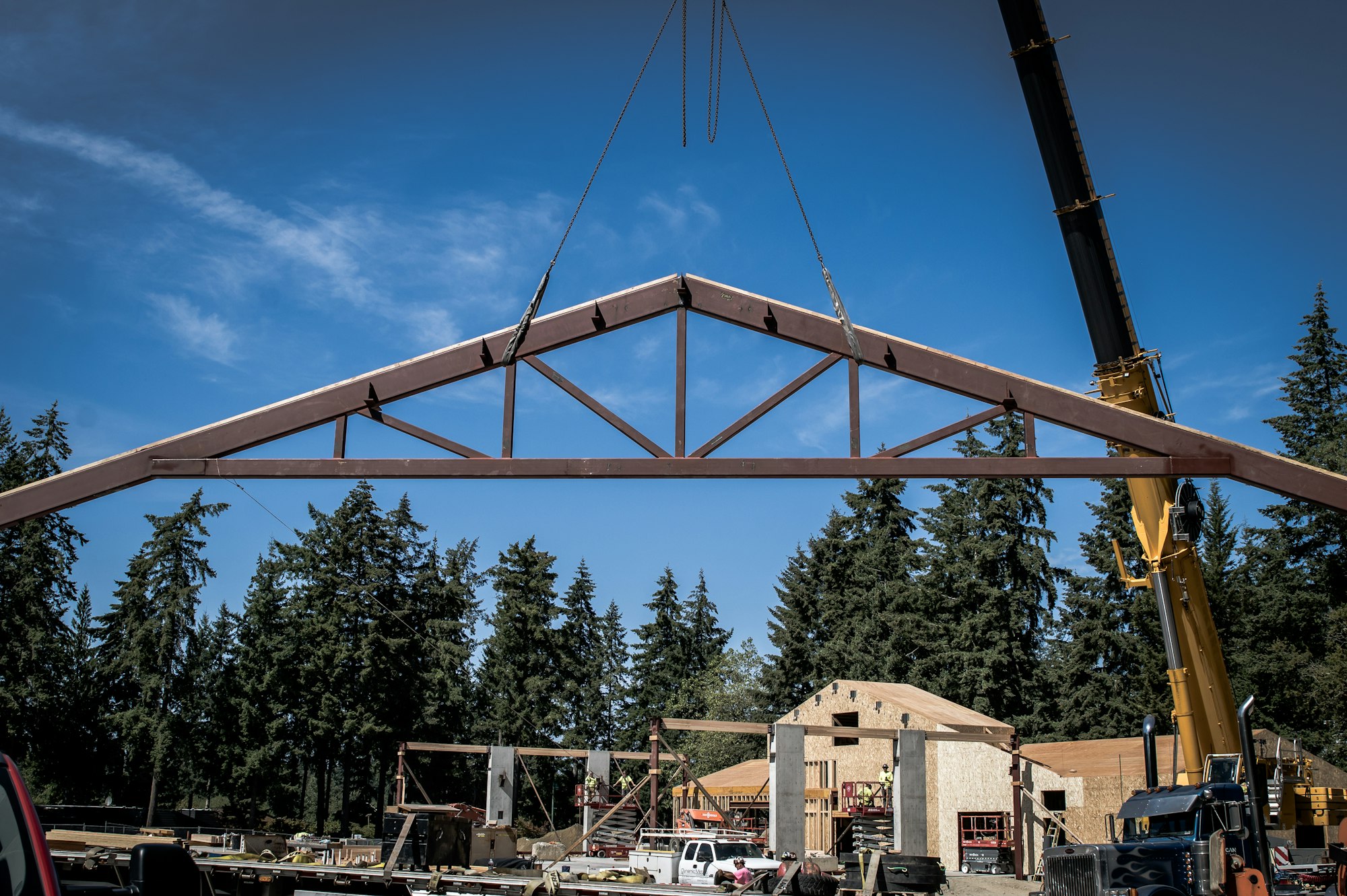 A crane lifts a truss frame at a construction site surrounded by trees and buildings under construction against a blue sky.