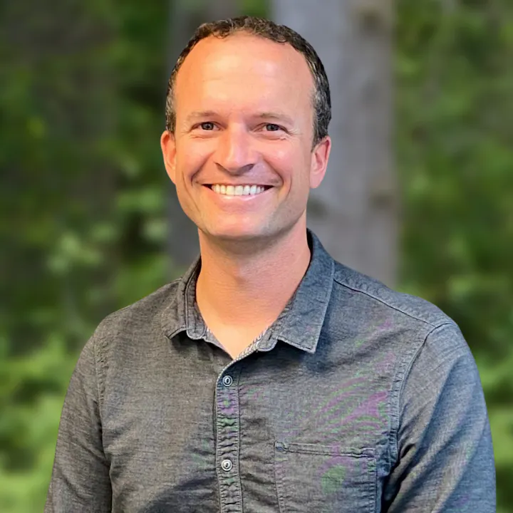 A smiling person with short hair wearing a button-up shirt stands against a blurred green background.