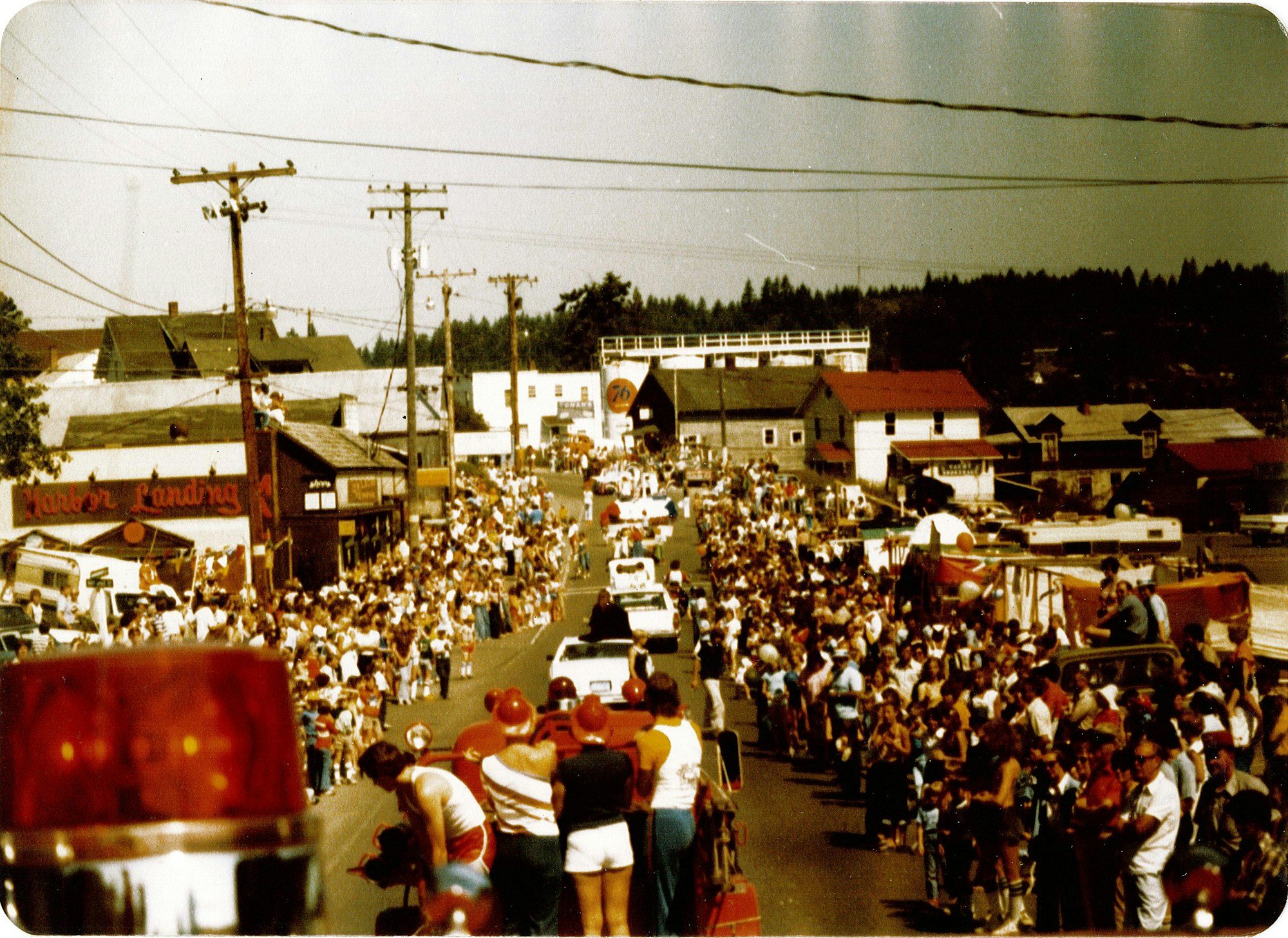 A vintage parade scene with a large crowd lining the street, various vehicles, and shops in a small town setting.