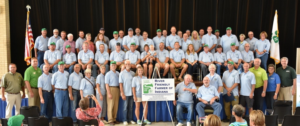 A large group of people posing for a photo, some wearing green caps, with a "River Friendly Farmer of Indiana" sign.