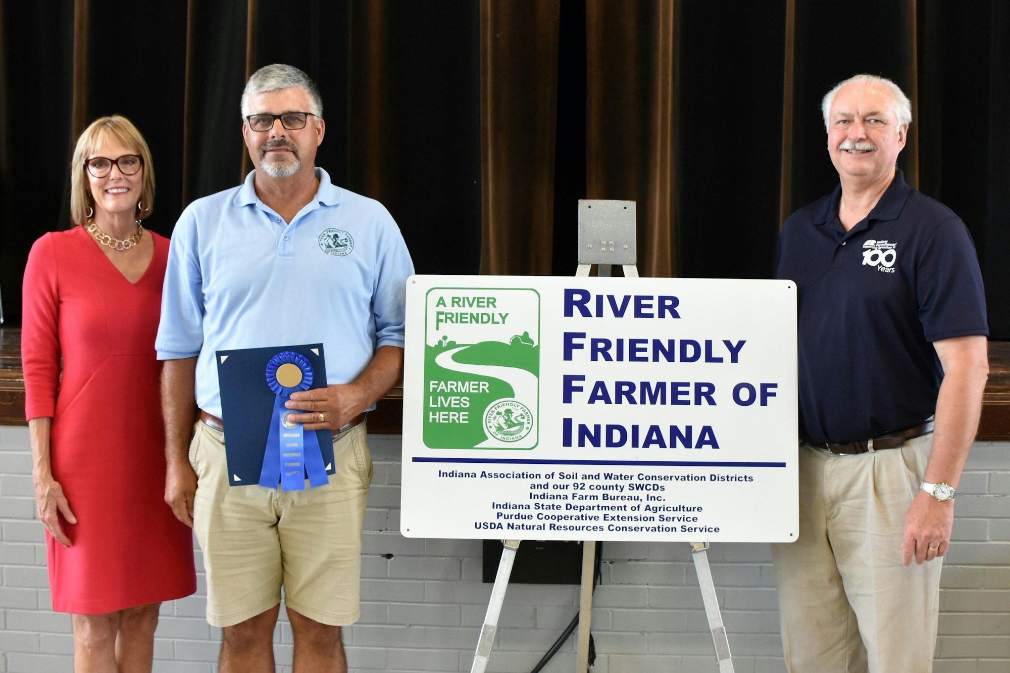 Three people stand beside a "River Friendly Farmer of Indiana" sign and an award ribbon.