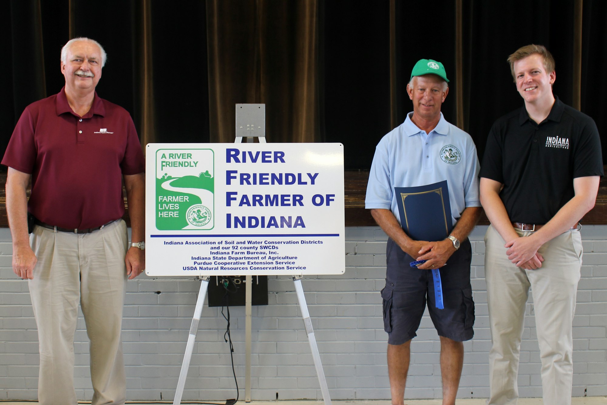 Three men stand beside a "River Friendly Farmer of Indiana" sign, one holding a certificate.