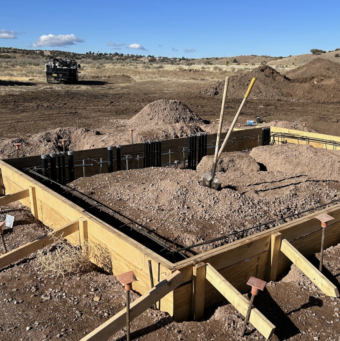 A construction site with wooden forms for concrete, steel rebar, tools, and a dump truck in the background.
