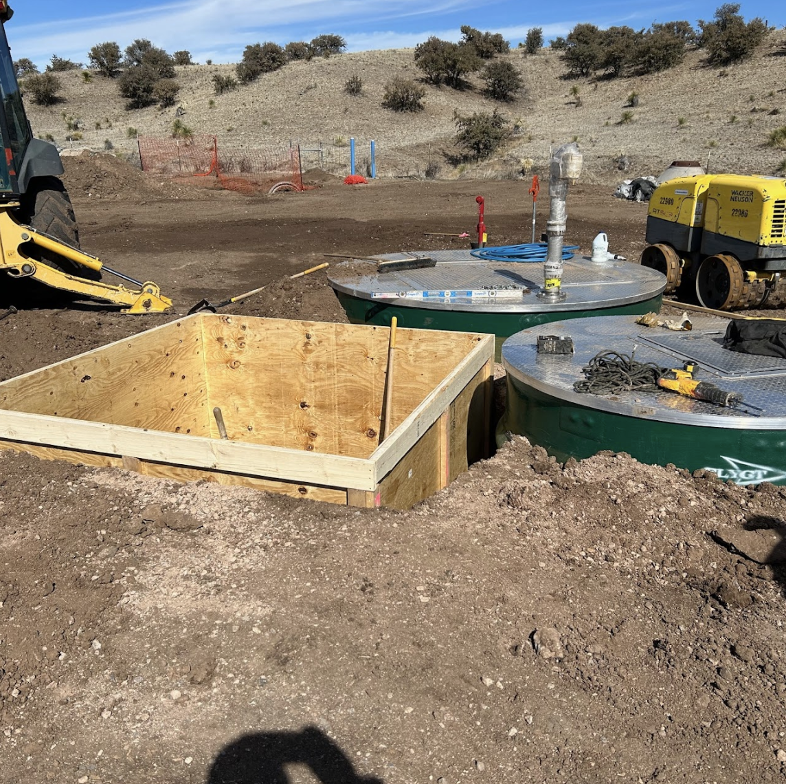 Construction site with wooden forms and septic tanks, excavator in the background.