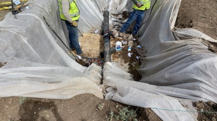 Workers in hi-vis vests at an excavation site with a pipe, geotextile fabric, and scattered materials.