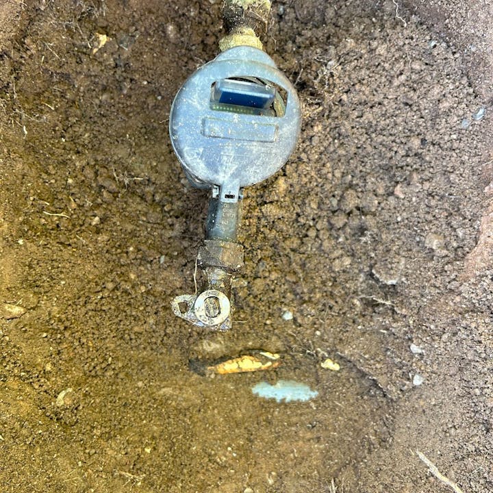A water meter and valve partially buried in soil, with slight water leakage evident from wet ground.