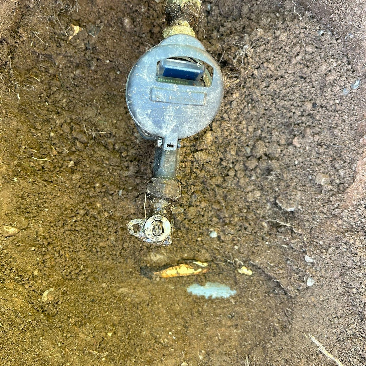 A water meter and valve partially buried in soil, with slight water leakage evident from wet ground.
