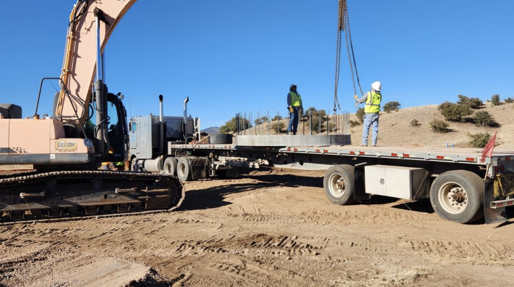 An excavator, two people in safety gear on a flatbed trailer, and construction materials in an outdoor setting.