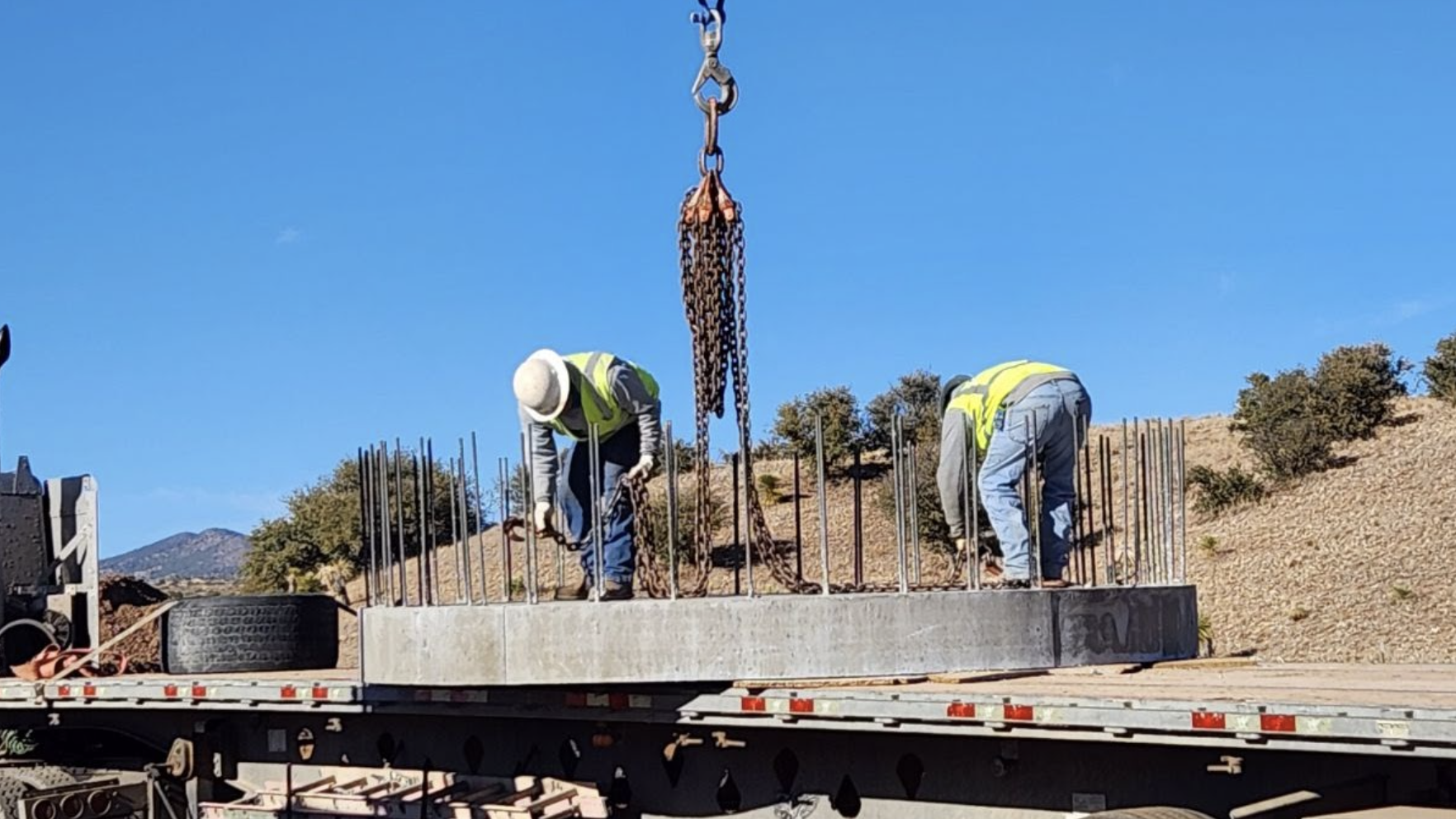Two workers in high-visibility vests are adjusting chains on a concrete slab being lifted by a crane onto a flatbed truck.