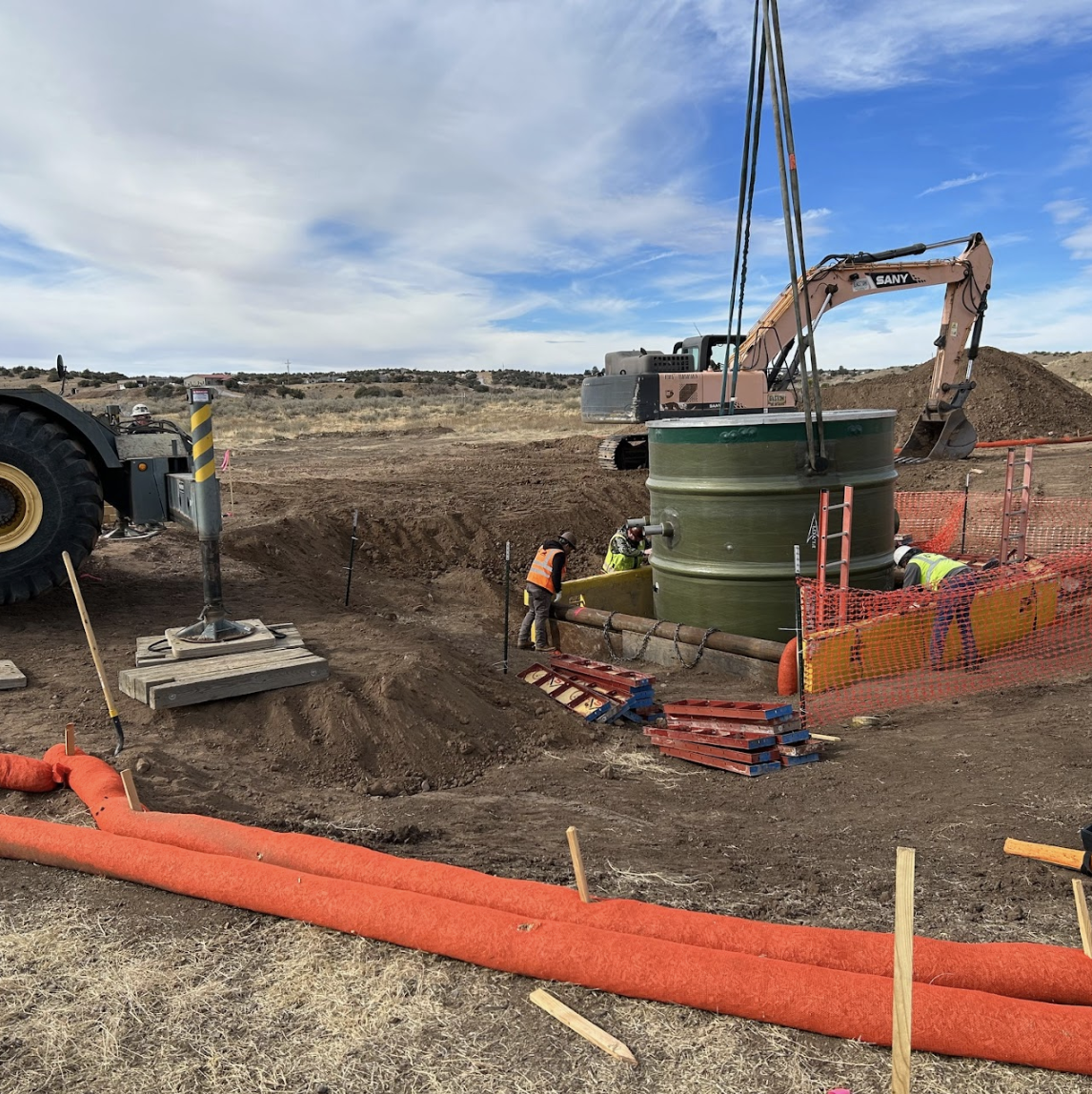 Construction site with an excavator, workers, and a large green pipe being installed in a trench.