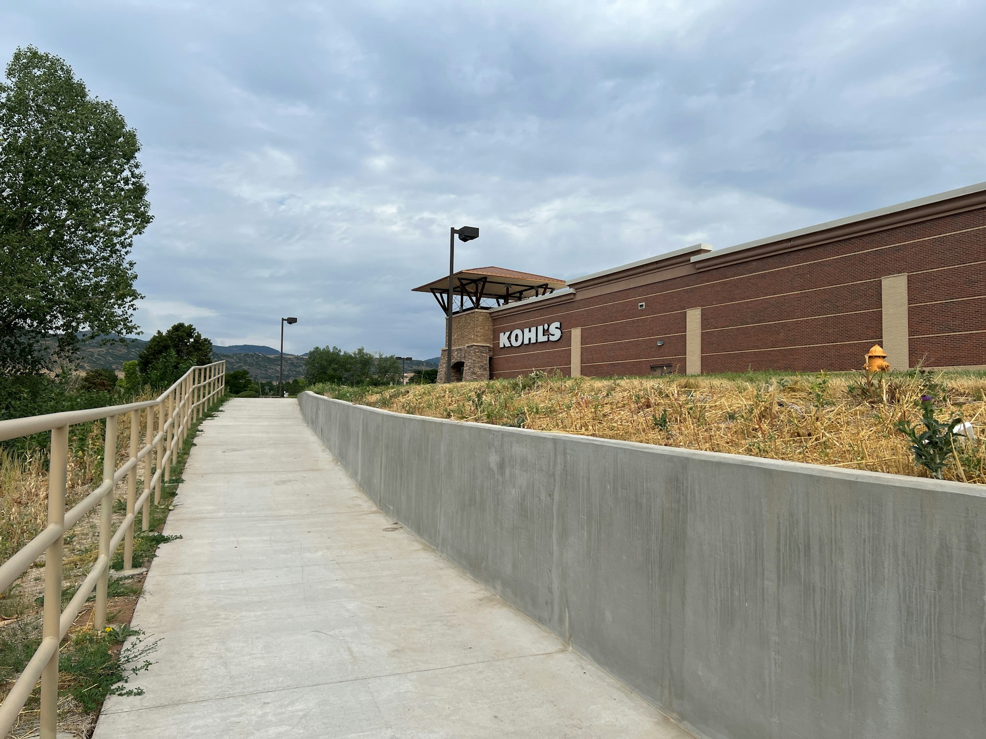 A walkway leads to a building with a "Kohl's" sign. It's bordered by a fence and grassy area under a cloudy sky.