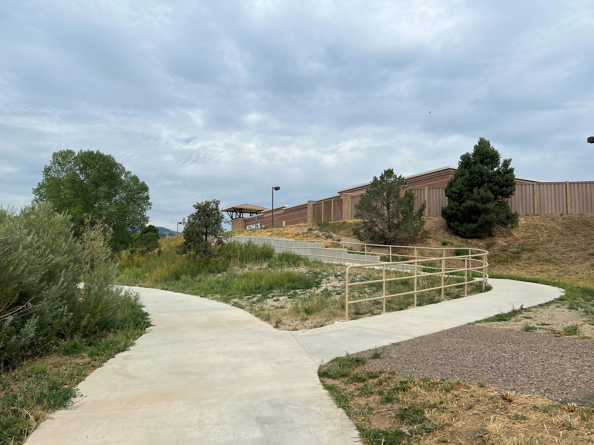 A curved concrete path with railings, surrounded by grass and trees, with a building and a cloudy sky in the background.