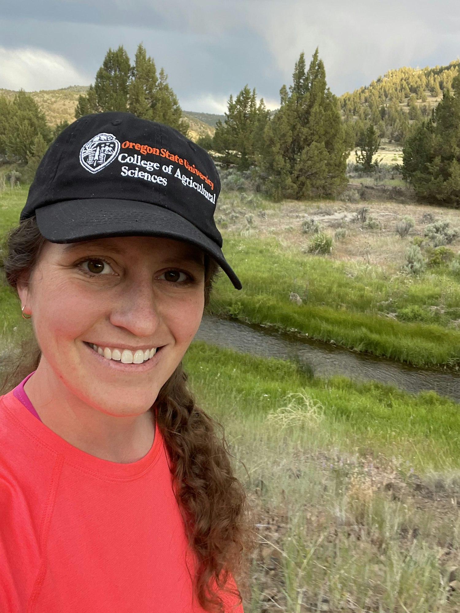 A person in a red shirt and cap is smiling outdoors near a grassy stream with trees and hills in the background.
