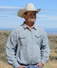 Man in a checkered shirt and cowboy hat standing outdoors with hills and blue sky in the background.