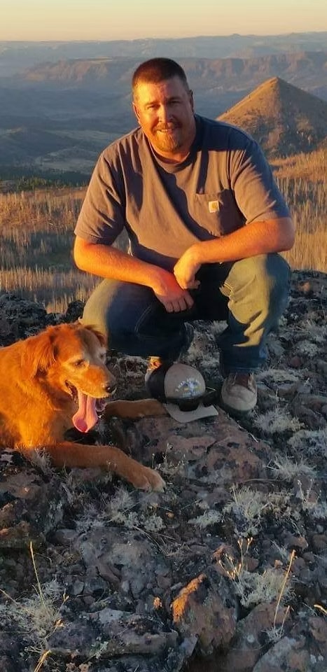 A man crouches on rocky terrain with a dog beside him, overlooking a mountainous landscape.