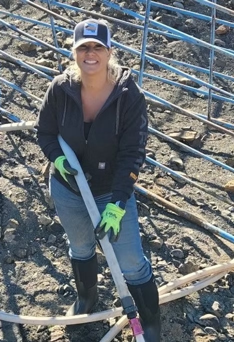A person in outdoor work clothing holding a large hose on a rocky terrain with blue tubing around.