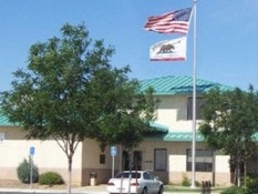 A building with teal roof, two flags (US and California), a tree, and a white car in front.