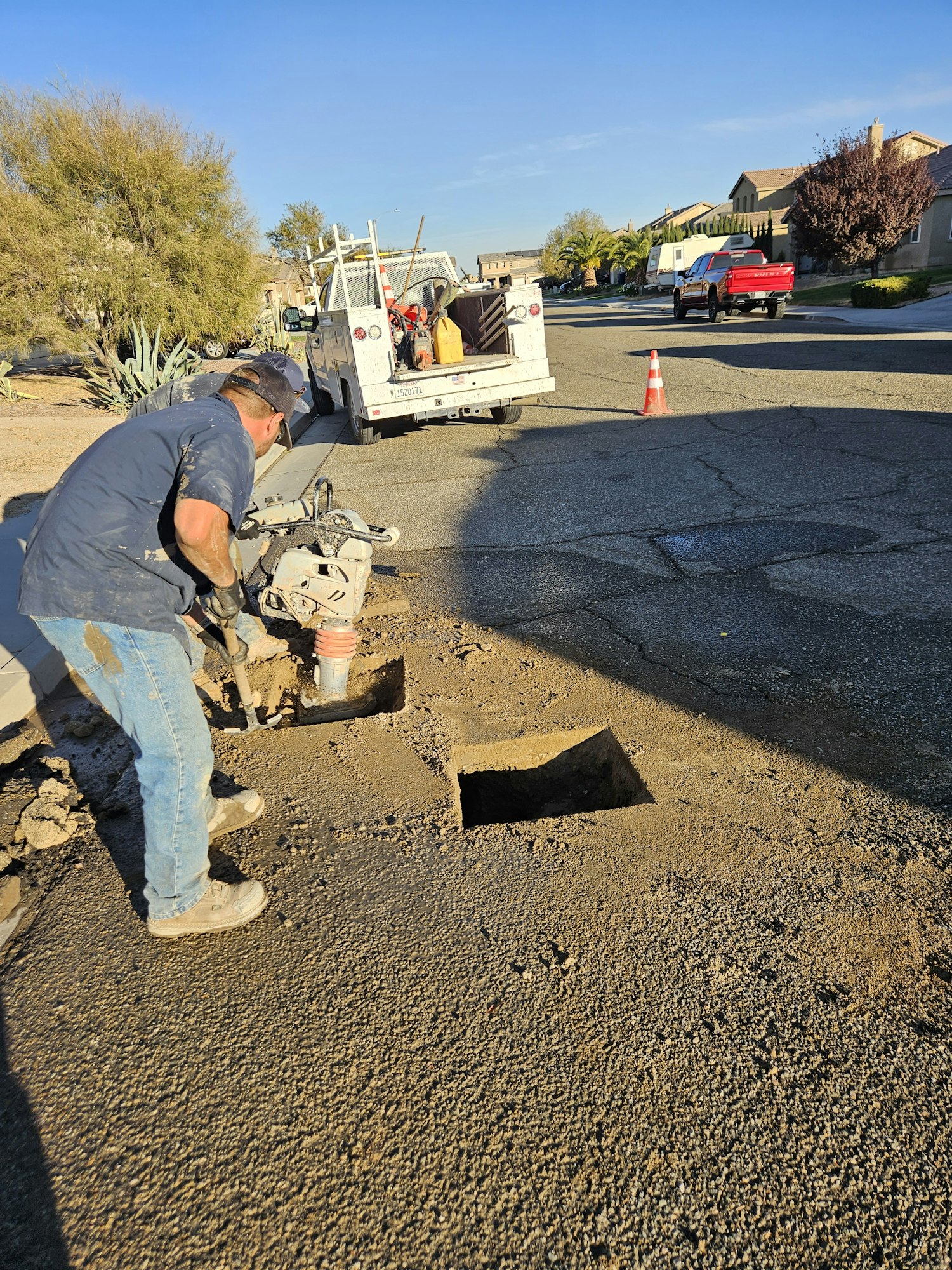 A worker operates machinery next to a square hole in the road; a utility truck is parked nearby.