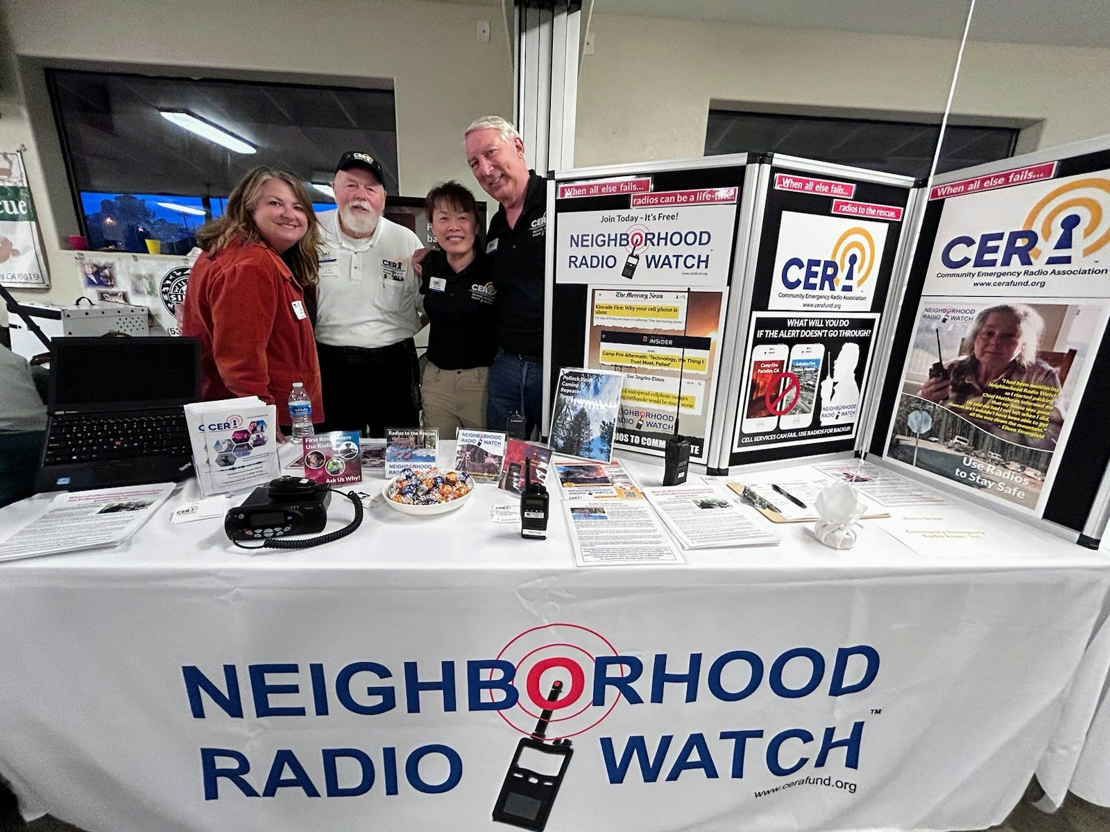 Four people representing the Community Emergency Radio Association and their 'Neighborhood Radio Watch' booth with information displays, a laptop, phone, and radios.