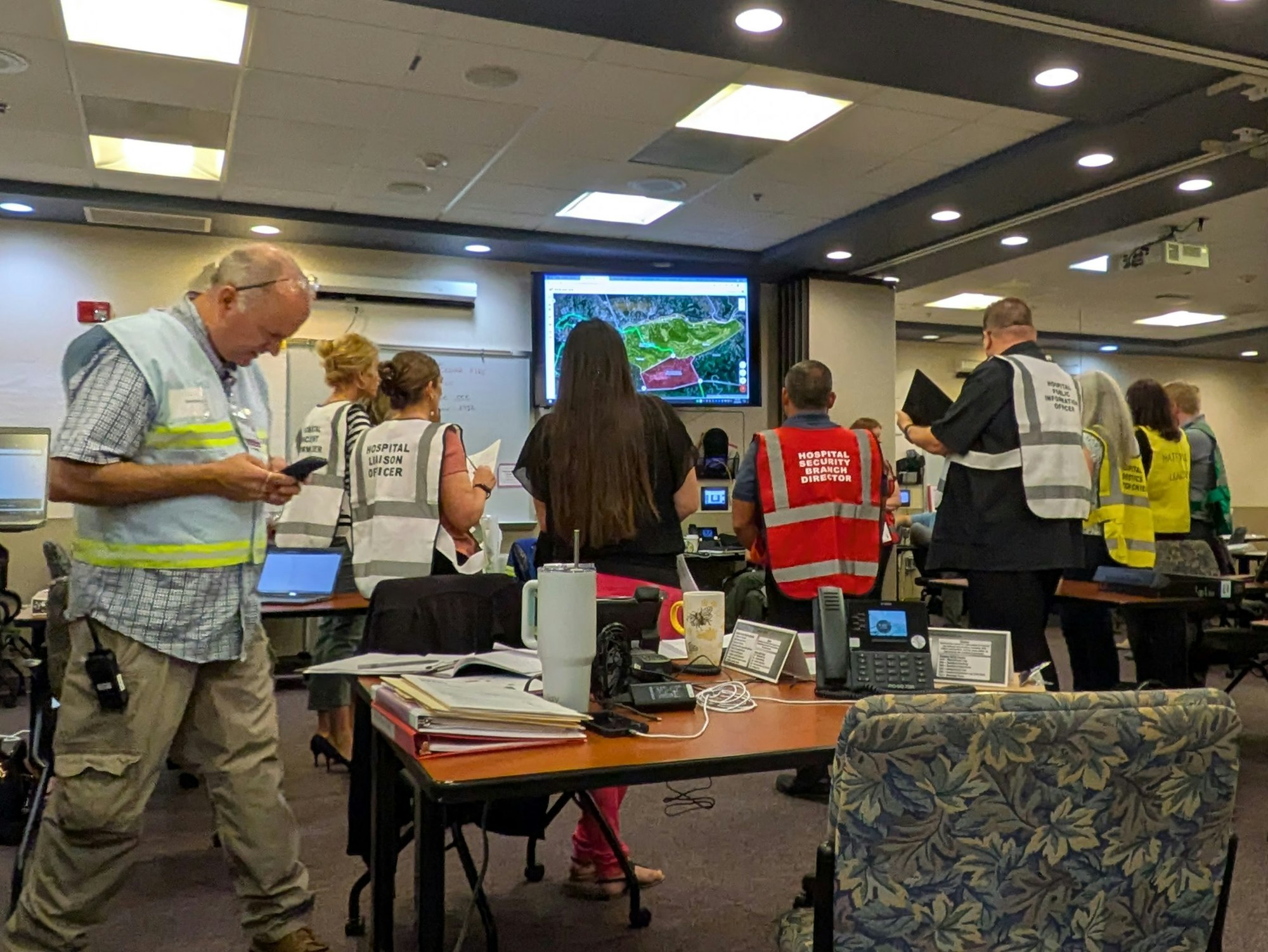 A group of people in vests are gathered in a room with maps displayed on screens, likely planning or coordinating an activity.