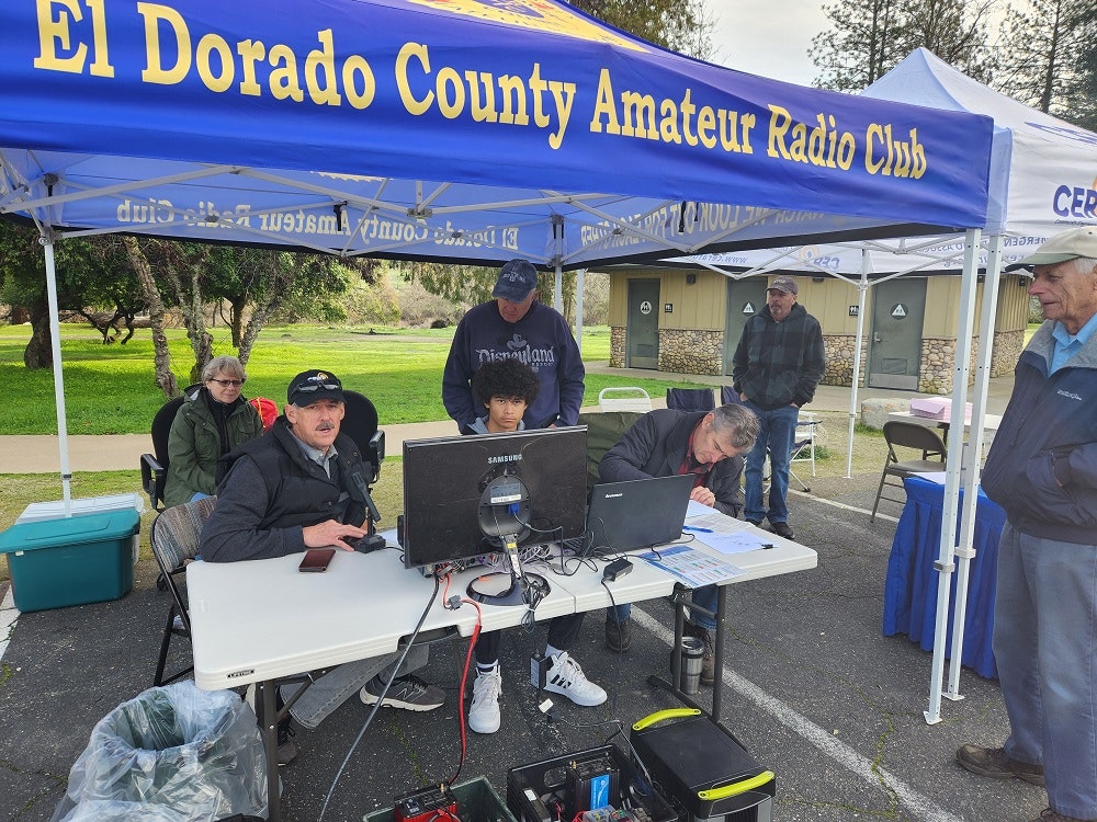 El Dorado County Amateur Radio Club enthusiasts gather under a club tent with equipment, tables, and chairs in a park-like setting for Winter Field Day 2024.