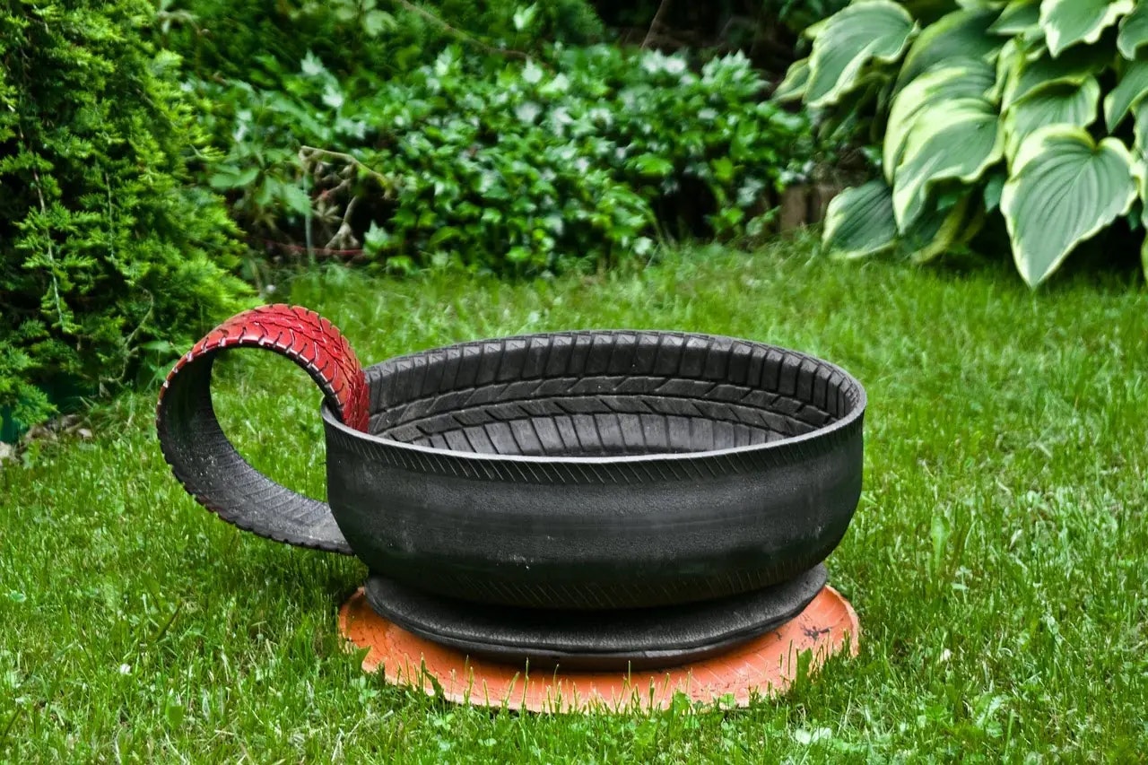 A large cup sculpture made from recycled tires with a red handle, placed on a grassy lawn near green plants.