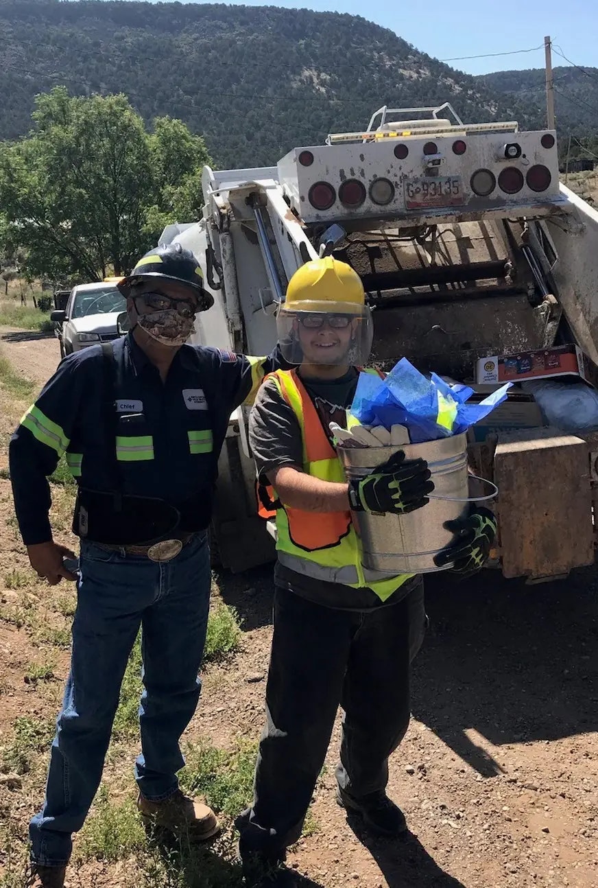 Two workers in safety gear posing by a garbage truck, one holding a bucket with blue tissue paper.