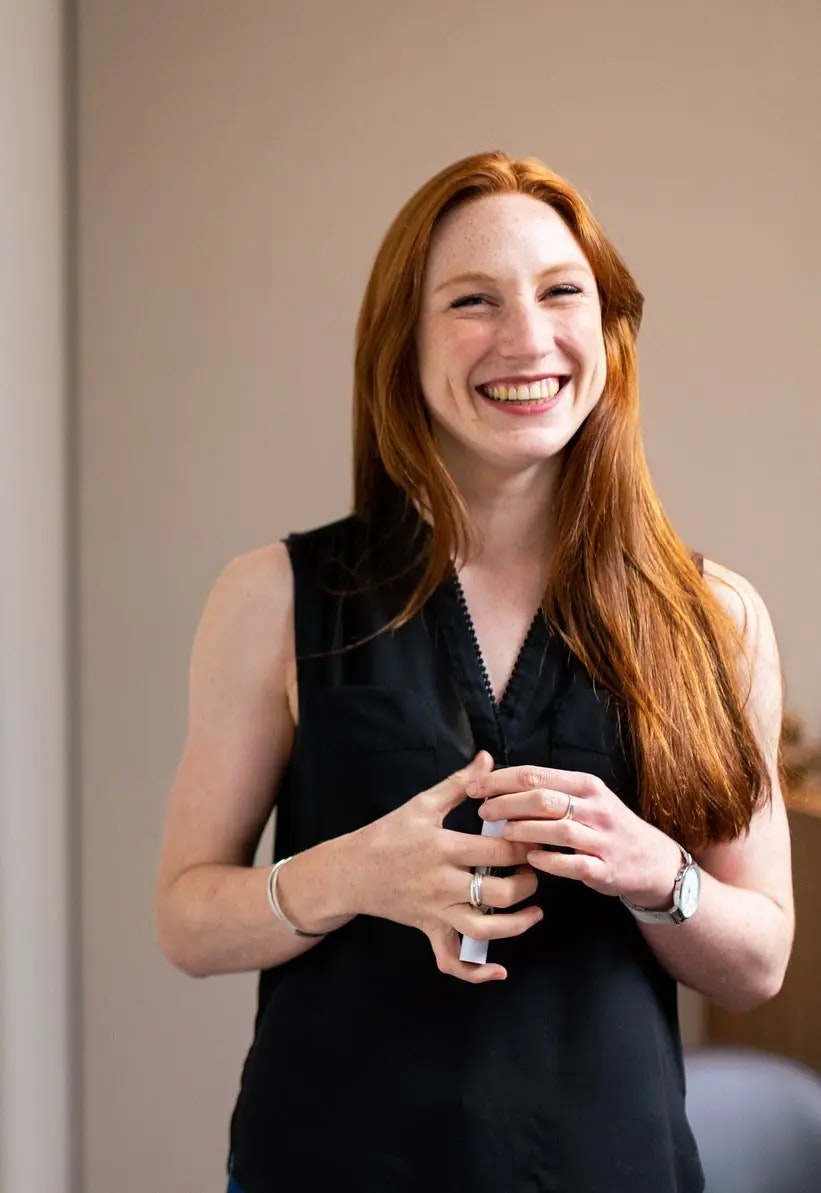 Smiling person with long red hair, wearing a sleeveless black top, posing indoors.