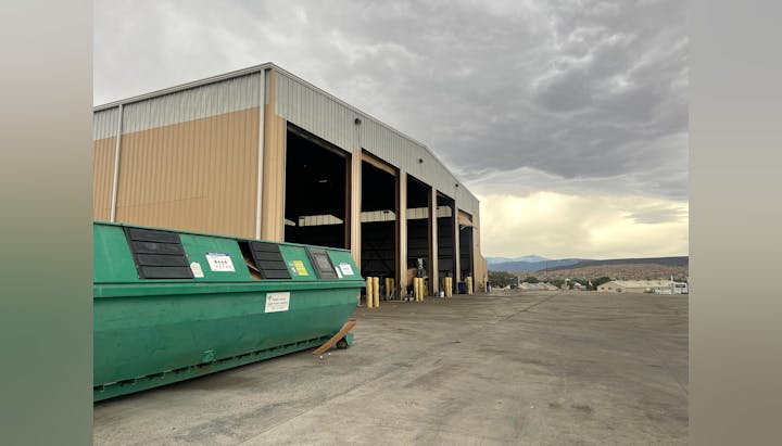 A green dumpster is placed outside a large building, with cloudy skies above and mountains in the background.