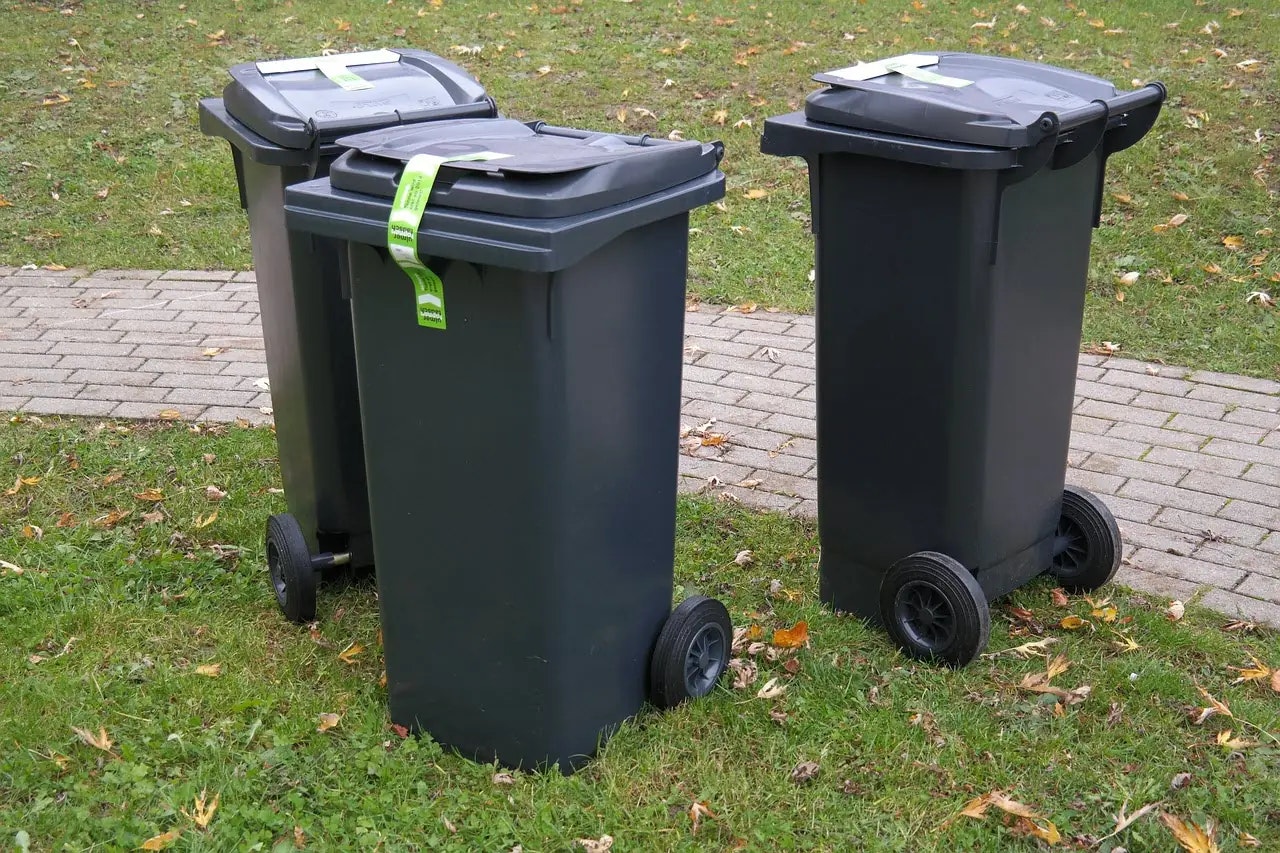 Three black wheeled garbage bins with green tags on a grass and paved area.
