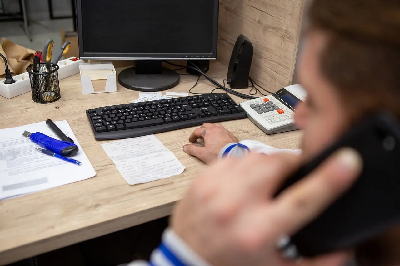 Office desk with a computer, keyboard, calculator, papers, and a person on the phone.