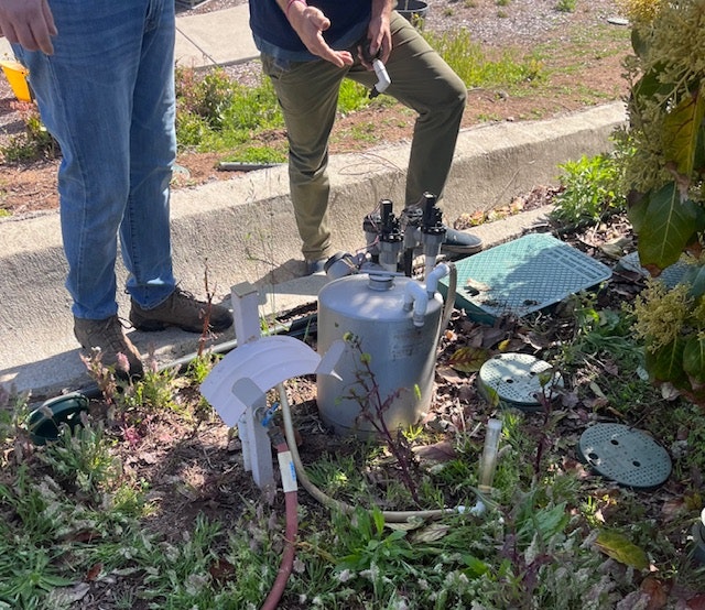 An outdoor setting with two people standing near a mechanical device and underground utilities exposed.