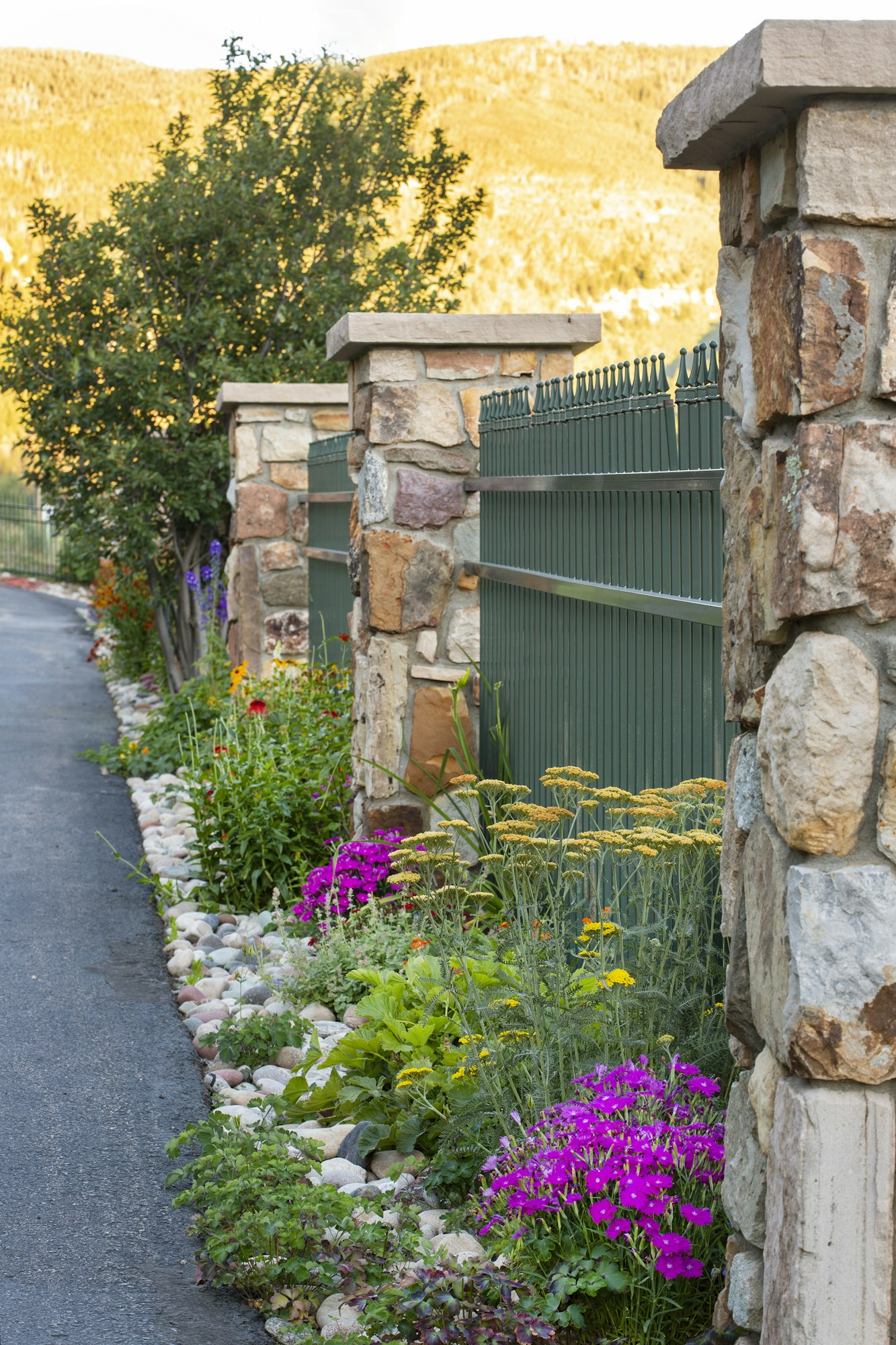 Stone fence, colorful flowers, pebbled garden path, and trees with mountains in the background.