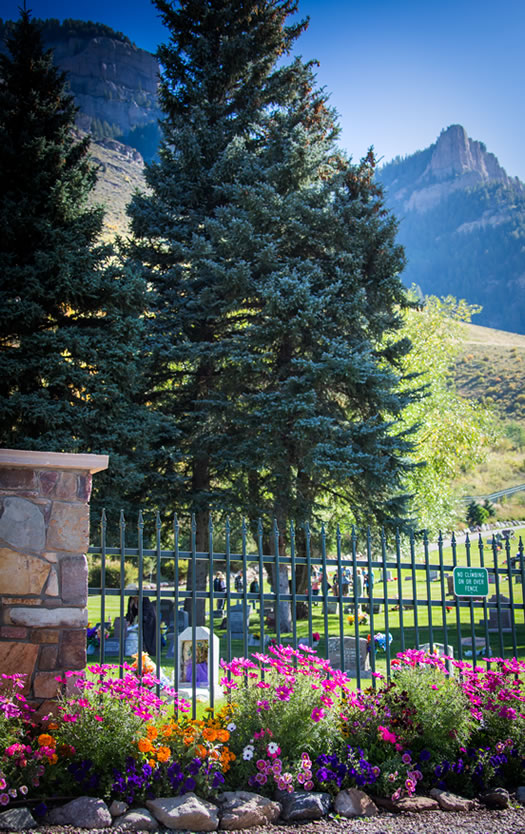 A cemetery with colorful flowers, trees, a fence, and mountain scenery in the background under a clear blue sky.