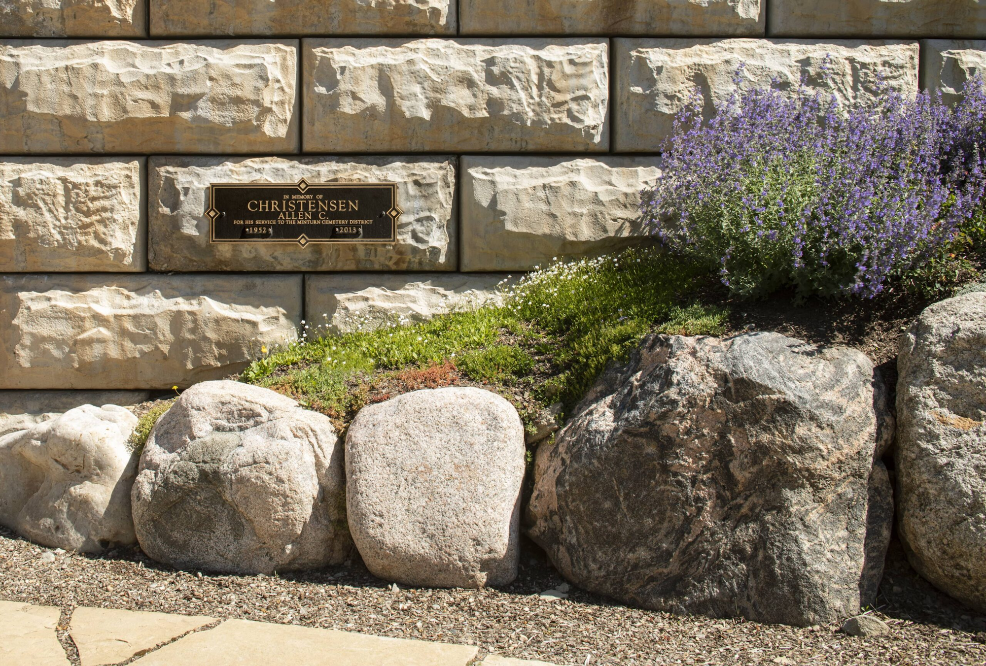 Stone wall with an in-memory plaque for Allen Christensen, surrounded by rocks, green grass, and purple flowers.